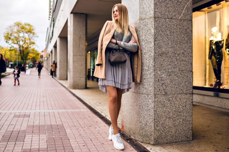 outdoor lifestyle fashion portrait pretty elegant glamour blonde woman with long legs wearing trendy sneakers dress sweater coat posing europe city traveling alone 1